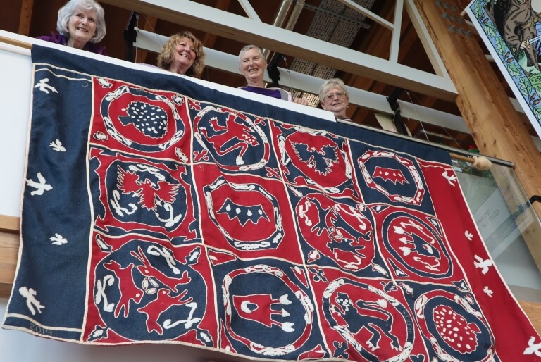 A blue and red piece of material with whit, blue and red symbols sewn on to a series of squares. The symbols depict crowns, a quiver with arrows, lions, eagles and three hares to symbolise Sherwood Forests' place as a former royal hunting forest. Four people are standing above it looking down over it.